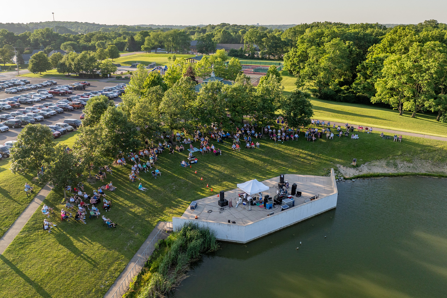 A large crowd gathers on a lawn by the lake for an outdoor summer concert at the park.
