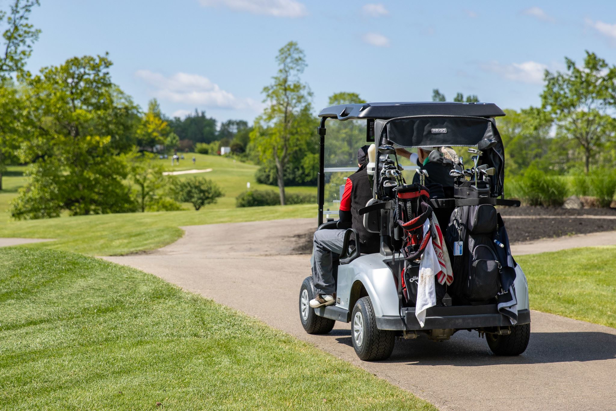 A golf cart carrying two golfers and clubs drives along a paved path toward distant fairways and tre
